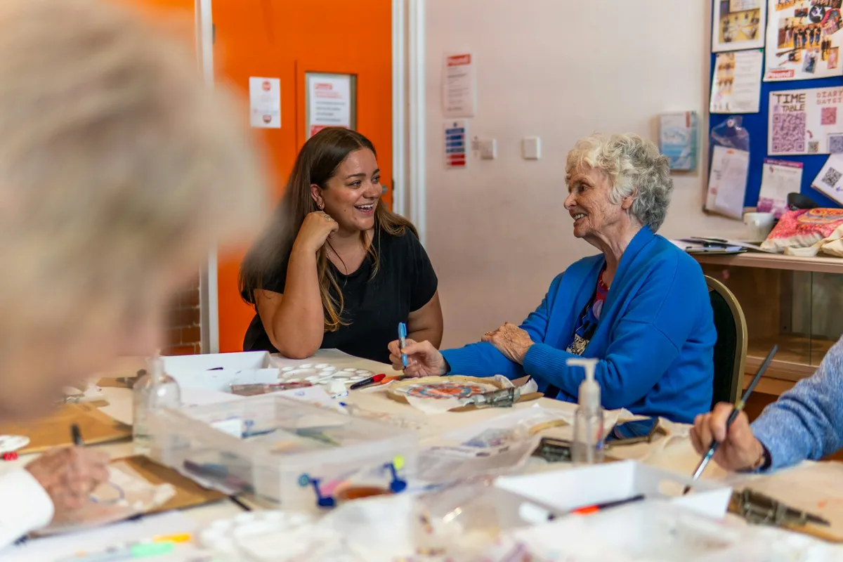 A smiling community worker chats with an older woman during an art activity at a community center